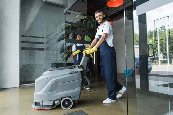 Homem feliz com máquina de limpeza de piso olhando para a câmera perto de mulher bi-racial limpeza porta de vidro — Fotografia de Stock