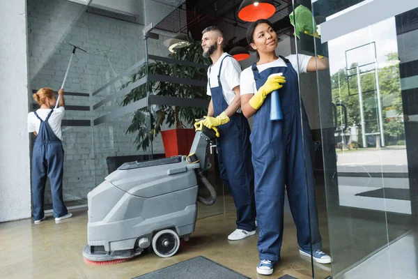 Lächelnder Mann mit Fußbodenwaschmaschine in der Nähe multiethnischer Frauen, die Glaseingänge von Büros reinigen — Stockfoto