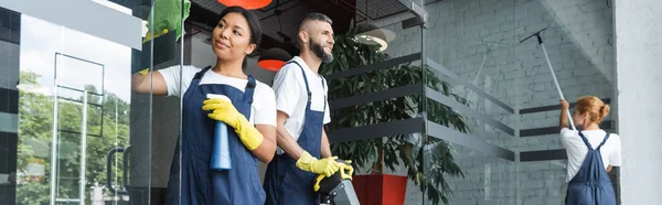 Mujeres interraciales lavando la entrada de cristal en la oficina cerca del hombre con la máquina de fregar piso, pancarta - foto de stock