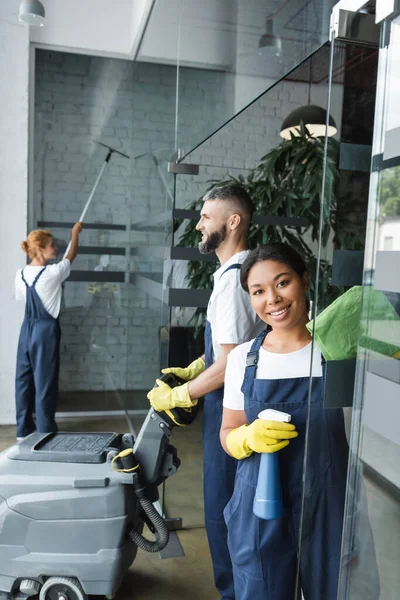 Mujer bi-racial sonriente con detergente mirando a la cámara cerca de colegas de limpieza de la oficina - foto de stock