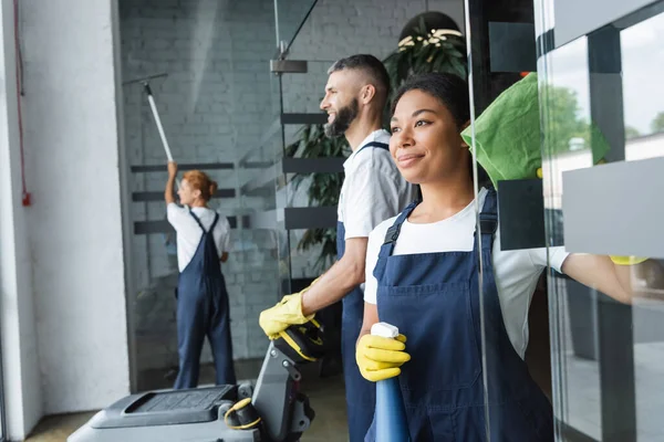 Happy bi-racial woman with rag and spray bottle near colleagues working in office — Stock Photo
