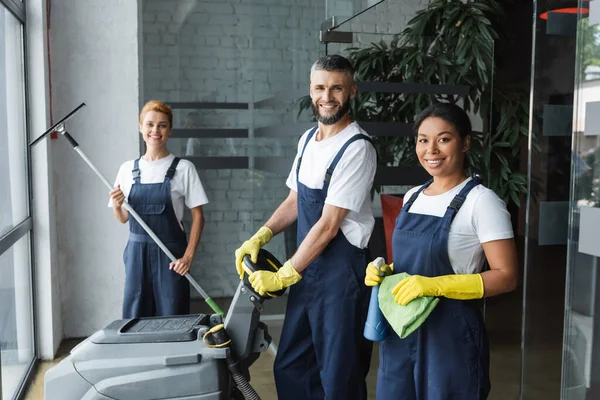 Equipo interracial feliz de limpiadores profesionales con suministros de limpieza mirando a la cámara en la oficina - foto de stock