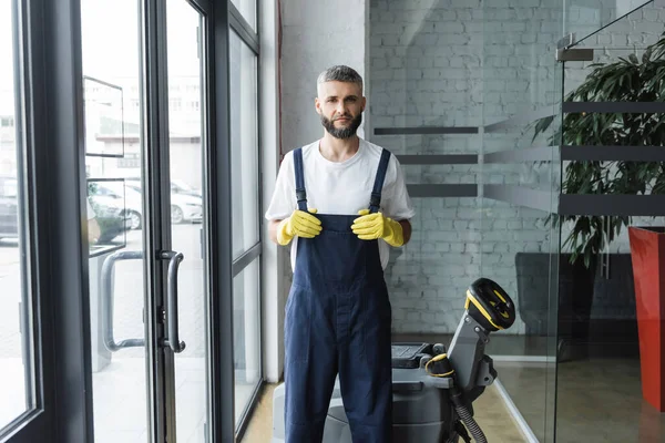 Бородатий чоловік в комбінезоні і гумові рукавички дивиться на камеру біля підлогового скраб машини — стокове фото