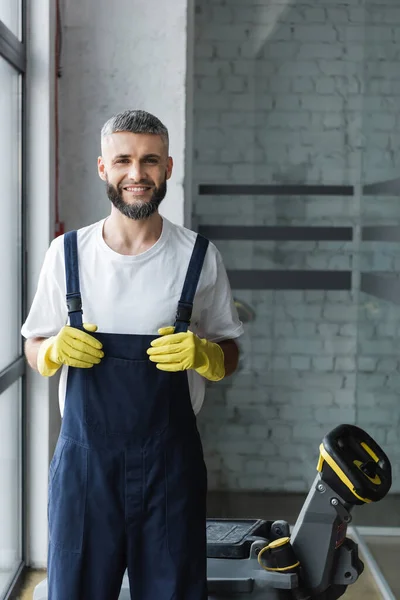 Cheerful man in rubber gloves and overalls smiling at camera near electrical floor scrubber machine — Stock Photo