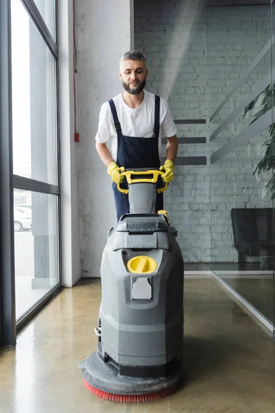 Hombre barbudo en overoles piso de limpieza con máquina de fregar eléctrica - foto de stock