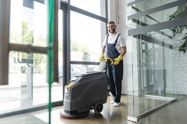 Full length of professional cleaner with electrical floor scrubber machine smiling at camera — Stock Photo