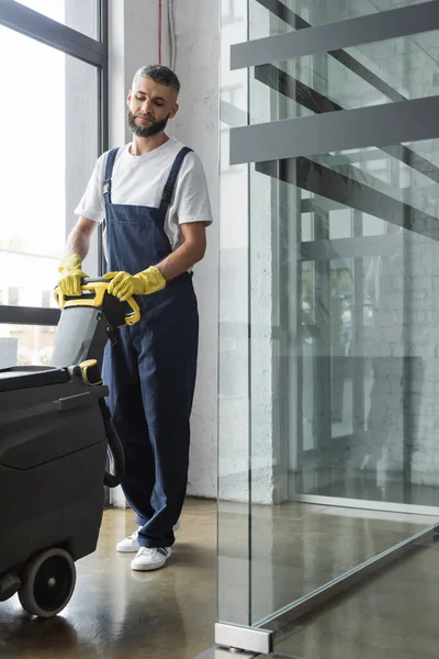 Longitud completa del hombre en overoles oficina de limpieza con máquina de fregar piso eléctrico - foto de stock