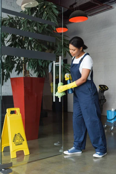 Longitud completa de la mujer bi-racial en uniforme lavando la puerta de cristal con el trapo en la oficina - foto de stock