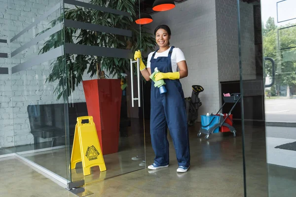 Limpador bi-racial alegre com pano e detergente olhando para a câmera perto da porta de vidro — Fotografia de Stock