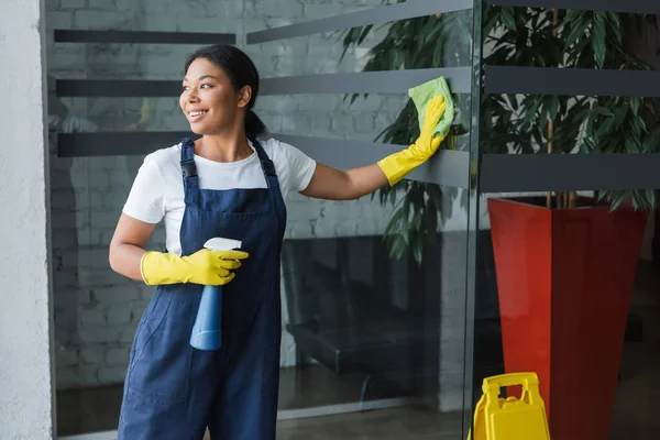 Felice donna bi-razziale guardando altrove e asciugando porta di vetro in ufficio — Foto stock