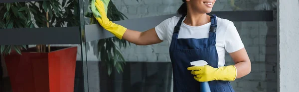Cropped view of smiling bi-racial woman in rubber gloves cleaning glass door in office, banner — Stock Photo
