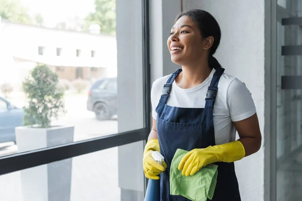 Mulher bi-racial alegre com pano e detergente olhando através da janela no escritório — Fotografia de Stock