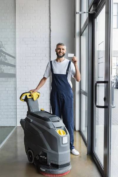 Comprimento total do limpador feliz mostrando cartão de visita em branco perto da máquina de limpeza de piso — Fotografia de Stock
