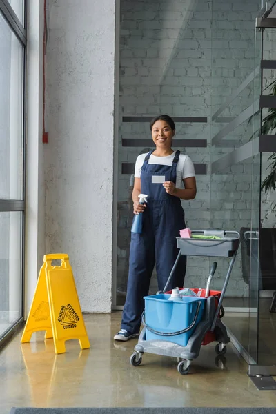 Feliz mulher bi-racial segurando detergente e vazio carrinho limpo cartão de visita com suprimentos de limpeza — Fotografia de Stock