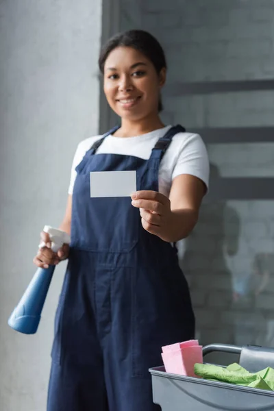 Mujer bi-racial borrosa con botella de spray que muestra la tarjeta de visita en blanco - foto de stock