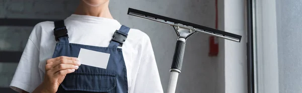 Cropped view of woman in overalls standing with window wiper and empty business card, banner — Stock Photo