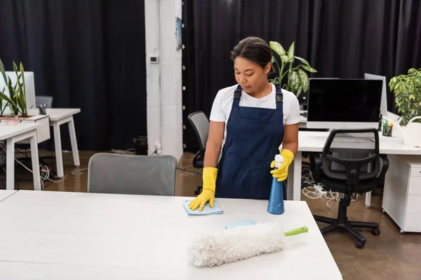 Young bi-racial cleaner in professional uniform washing desk in office — Stock Photo