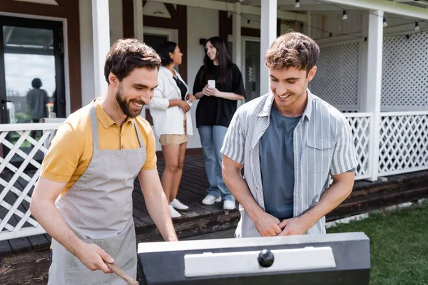 Des hommes souriants cuisinent près du gril et brouillent les amis interracial dans la cour arrière — Photo de stock