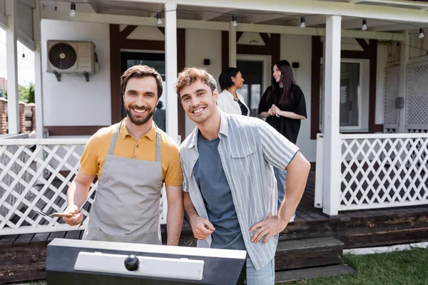 Smiling friends looking at camera near grill and blurred interracial women in backyard — Stock Photo