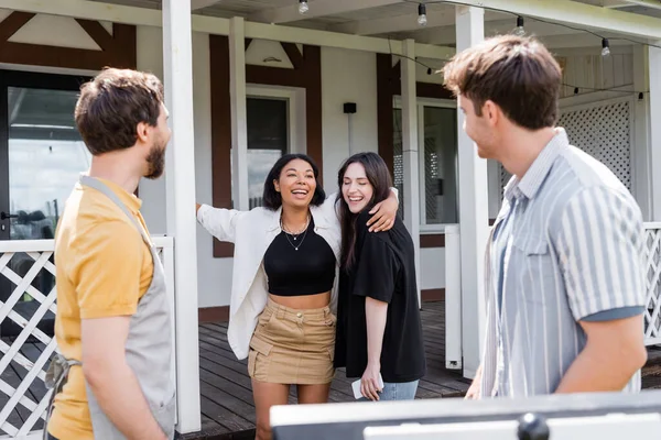 Mujeres interraciales abrazándose cerca de amigos y parrilla borrosa en el patio trasero - foto de stock