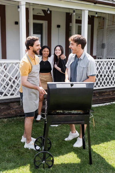 Cheerful multiethnic friends talking while cooking on grill in backyard — Stock Photo