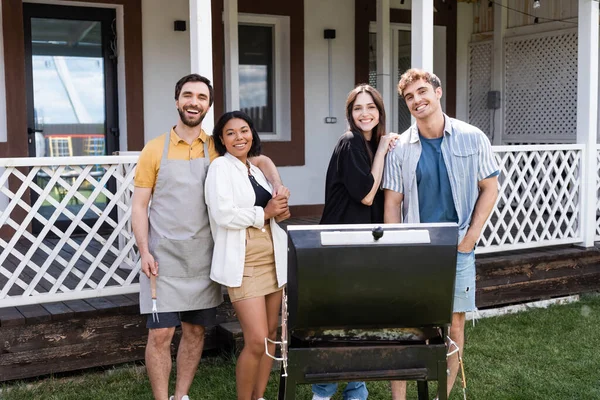 Positive interracial friends hugging and looking at camera near grill in backyard — Stock Photo
