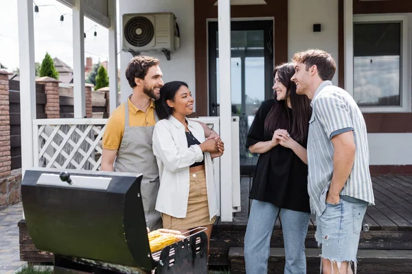 Couples multiethniques souriants parlant près du gril pendant le pique-nique dans la cour arrière — Photo de stock