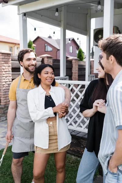 Hombre sonriente en delantal abrazando novia bi-racial cerca de amigos en el patio trasero - foto de stock