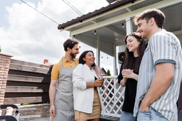 Cheerful interracial friends standing near sausages on grill during picnic — Stock Photo