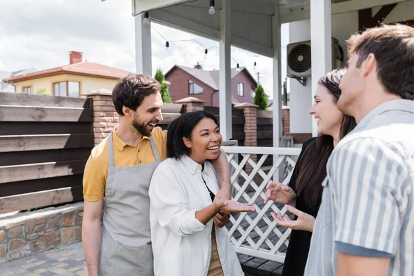 Mujer bi-racial hablando con amigos cerca de novio en delantal en el patio trasero - foto de stock