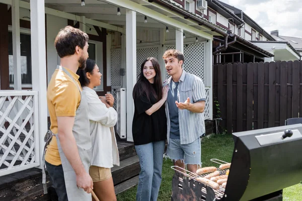 Casal positivo falando com amigos inter-raciais perto de churrasqueira ao ar livre — Fotografia de Stock