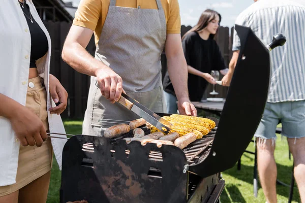 Man in apron cooking on grill near interracial friends in backyard — Stock Photo