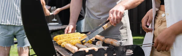 Cropped view of interracial friends cooking sausages and corn on grill in backyard, banner — Stock Photo