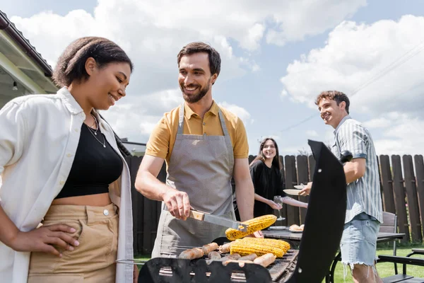 Uomo sorridente in grembiule cucina mais sulla griglia vicino amico bi-razziale in cortile — Foto stock