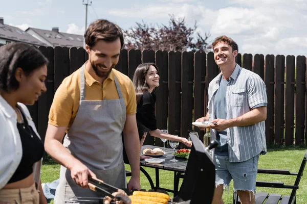 Sonriente hombre sosteniendo placa cerca borrosa interracial amigos cocinar en la parrilla en el patio trasero - foto de stock