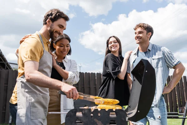 Uomo positivo in grembiule cucina mais alla griglia vicino bi-razziale fidanzata e amici in cortile — Foto stock