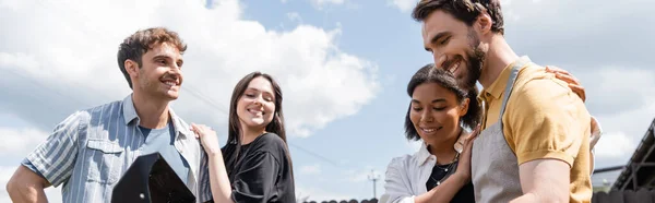 Smiling interracial couple looking at grill near friends outdoors, banner — Stock Photo