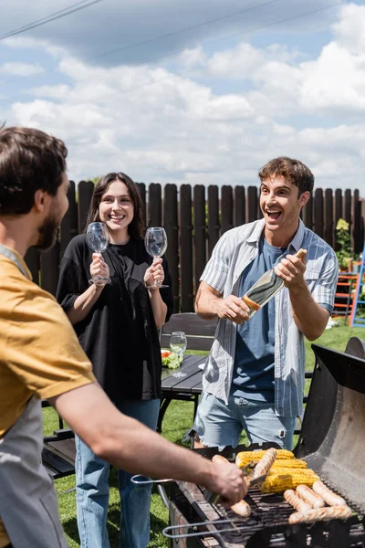 Cheerful man and woman holding wine and glasses near blurred friend cooking bbq in backyard — Stock Photo