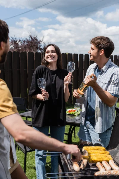 Smiling woman holding glasses near friends with wine and grill in backyard — Stock Photo