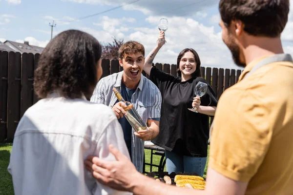 Allegro uomo e donna che tengono il vino vicino a amici multietnici offuscati e cibo sul barbecue in cortile — Foto stock