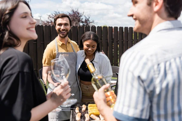 Sorrindo casal multiétnico abraçando perto bbq e amigos borrados com vinho ao ar livre — Fotografia de Stock