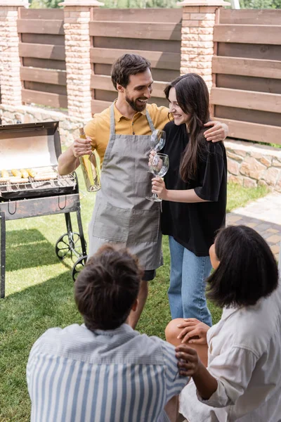 Uomo sorridente in grembiule abbracciando fidanzata e tenendo il vino vicino sfocato amici multietnici e grill all'aperto — Foto stock