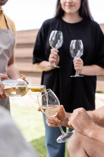 Обрізаний вид розмитого чоловіка в фартусі, що поливає вино поруч з друзями в окулярах на відкритому повітрі — стокове фото