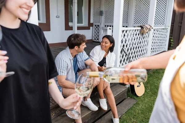 Multiethnic friends talking near acoustic guitar and blurred people with wine on backyard — Stock Photo