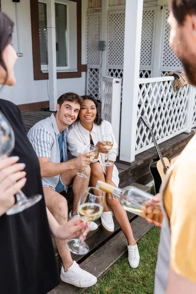 Smiling multiethnic friends holding wine near acoustic guitar and blurred people in backyard — Stock Photo