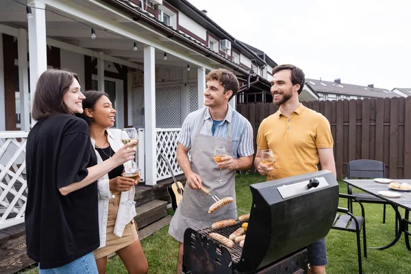 Positive men talking to interracial friends near grill during picnic with wine outdoors — Stock Photo