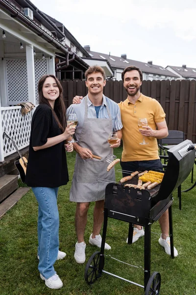 Smiling friends with glasses of wine looking at camera near food on grill in backyard — Stock Photo