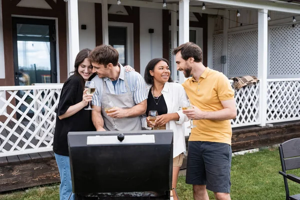 Sorrindo casais multiétnicos com vinho falando perto de churrasqueira no quintal — Fotografia de Stock