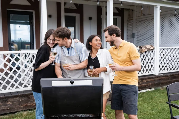 Couples multiethniques souriants avec du vin parlant près du gril dans la cour — Photo de stock