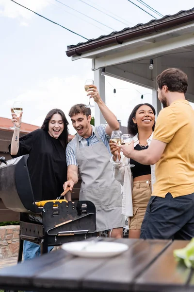 Emocionado hombre y mujer sosteniendo vino y cocinando en la parrilla mientras amigos interracial hablando en el patio trasero - foto de stock